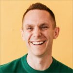 A person with short brown hair smiles broadly while looking slightly to the left. They are wearing a green sweater and stand against a solid yellow background.