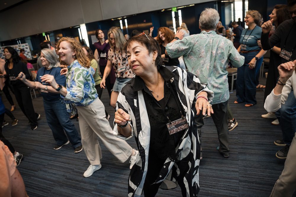 A group of people are dancing in a room. They are casually dressed, and some are smiling and laughing. The floor is carpeted and the walls have large windows, suggesting a spacious setting. A group of people are dancing in a room. They are casually dressed, and some are smiling and laughing. The floor is carpeted and the walls have large windows, suggesting a spacious setting.