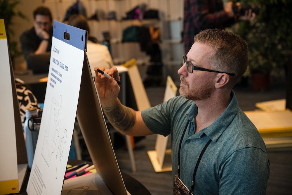 A person with glasses and a short-sleeve shirt is sitting and writing on a large notepad or easel in a conference or meeting setting. Other people are seen blurred in the background.