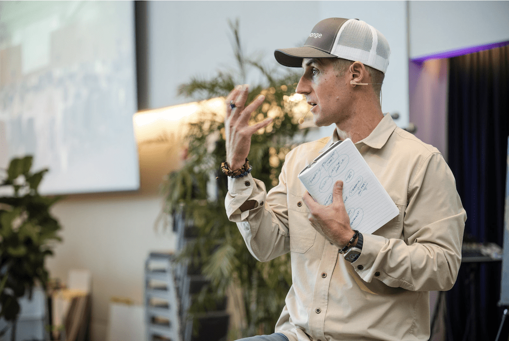 A person wearing a beige shirt and cap is speaking and gesturing with one hand while holding a notebook in the other. The background features a plant and a projection screen. A person wearing a beige shirt and cap is speaking and gesturing with one hand while holding a notebook in the other. The background features a plant and a projection screen.