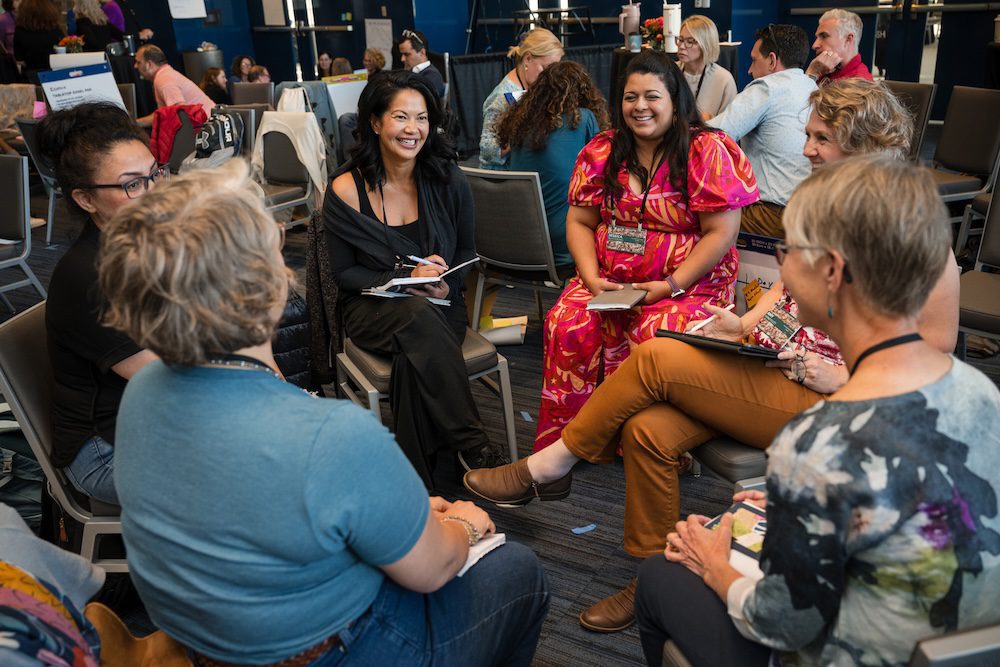 A group of people sit in a circle, engaged in conversation. They are seated in a conference or meeting room. Some hold notebooks and pens. The setting appears informal and relaxed.