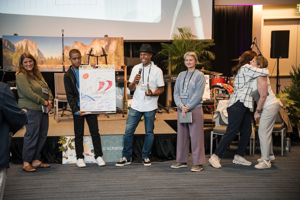 A group of people stands on a stage during a presentation. One person holds a large poster with drawings and signatures. In the background, a screen and musical instruments are visible. Two individuals hug on the right side of the stage.