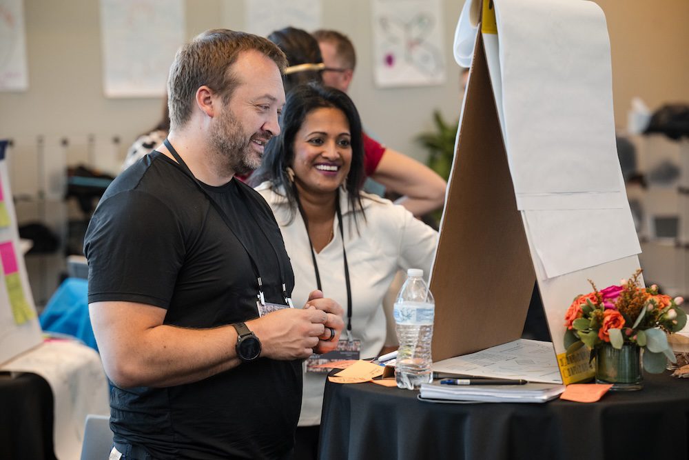 A man and woman engage in conversation at a table in a conference setting. They are looking at a display board with paper. There is a bottle of water and a small floral arrangement on the table. Other attendees and posters are in the background.