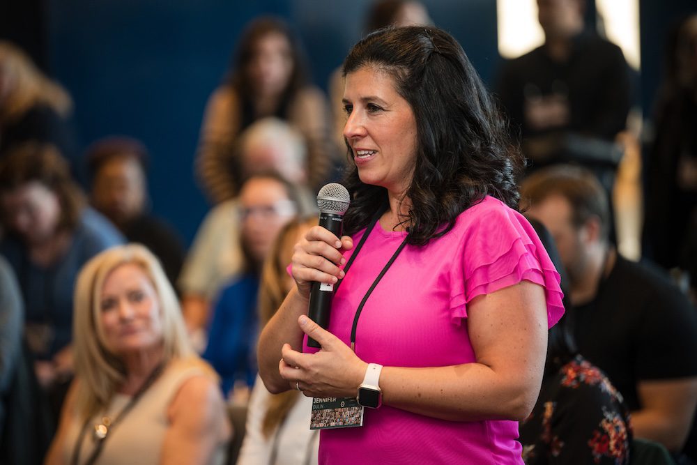 A woman in a pink blouse speaks into a microphone while standing in a room filled with people seated in the background. She wears a name tag and a smartwatch. The setting appears to be a conference or seminar.