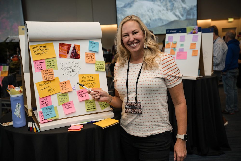 A woman stands smiling next to a large flip chart covered with colorful sticky notes. She is holding a pen, and a water bottle is visible on the table. Other people can be seen in the background, along with another board with notes.