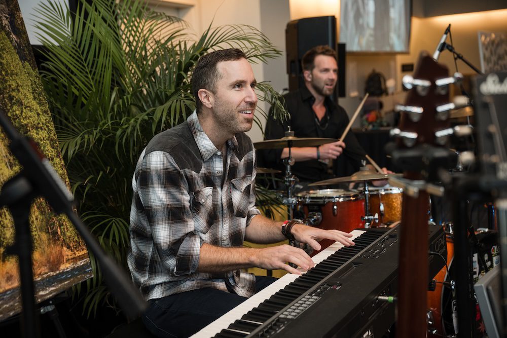 A smiling man plays a keyboard while another man plays drums in the background. They are performing indoors, with a leafy plant and a large painting visible behind them. Musical equipment is in the foreground. A smiling man plays a keyboard while another man plays drums in the background. They are performing indoors, with a leafy plant and a large painting visible behind them. Musical equipment is in the foreground.