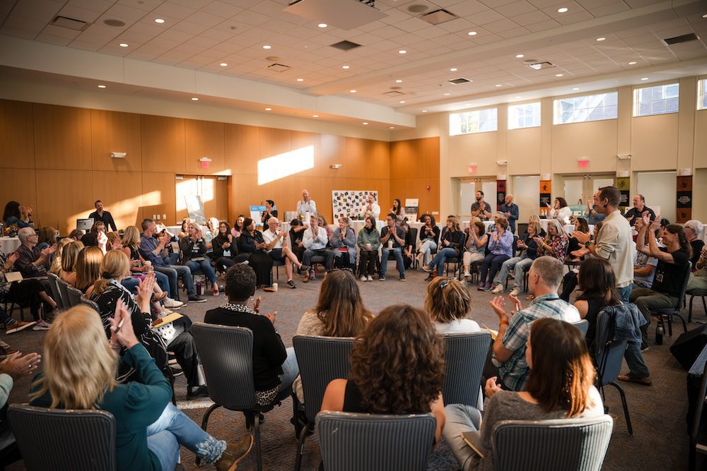 A group of people are seated in a large circle in a well-lit conference room. Some are clapping, and others are listening to a speaker at the front. The room has beige walls and large windows letting in natural light. A group of people are seated in a large circle in a well-lit conference room. Some are clapping, and others are listening to a speaker at the front. The room has beige walls and large windows letting in natural light.