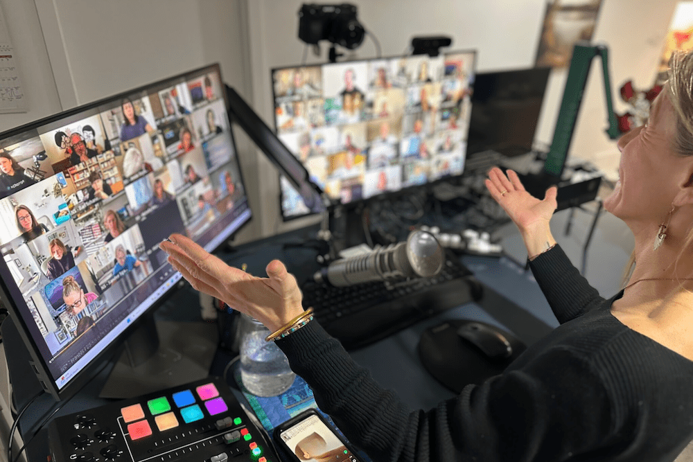 A woman sits at a desk with two monitors displaying a video conference with many participants. A microphone, audio mixer, water bottle, and phone are also on the desk. The woman gestures with open hands. A woman sits at a desk with two monitors displaying a video conference with many participants. A microphone, audio mixer, water bottle, and phone are also on the desk. The woman gestures with open hands.