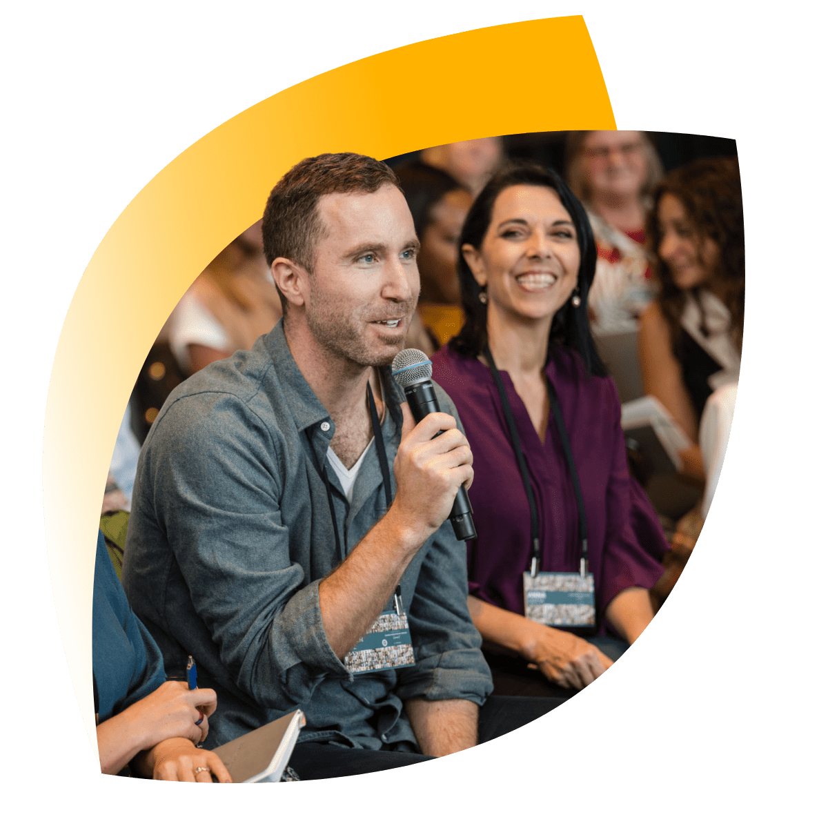 A man holds a microphone and speaks while seated among a group of people at an indoor event. Others around him listen and smile. A man holds a microphone and speaks while seated among a group of people at an indoor event. Others around him listen and smile.