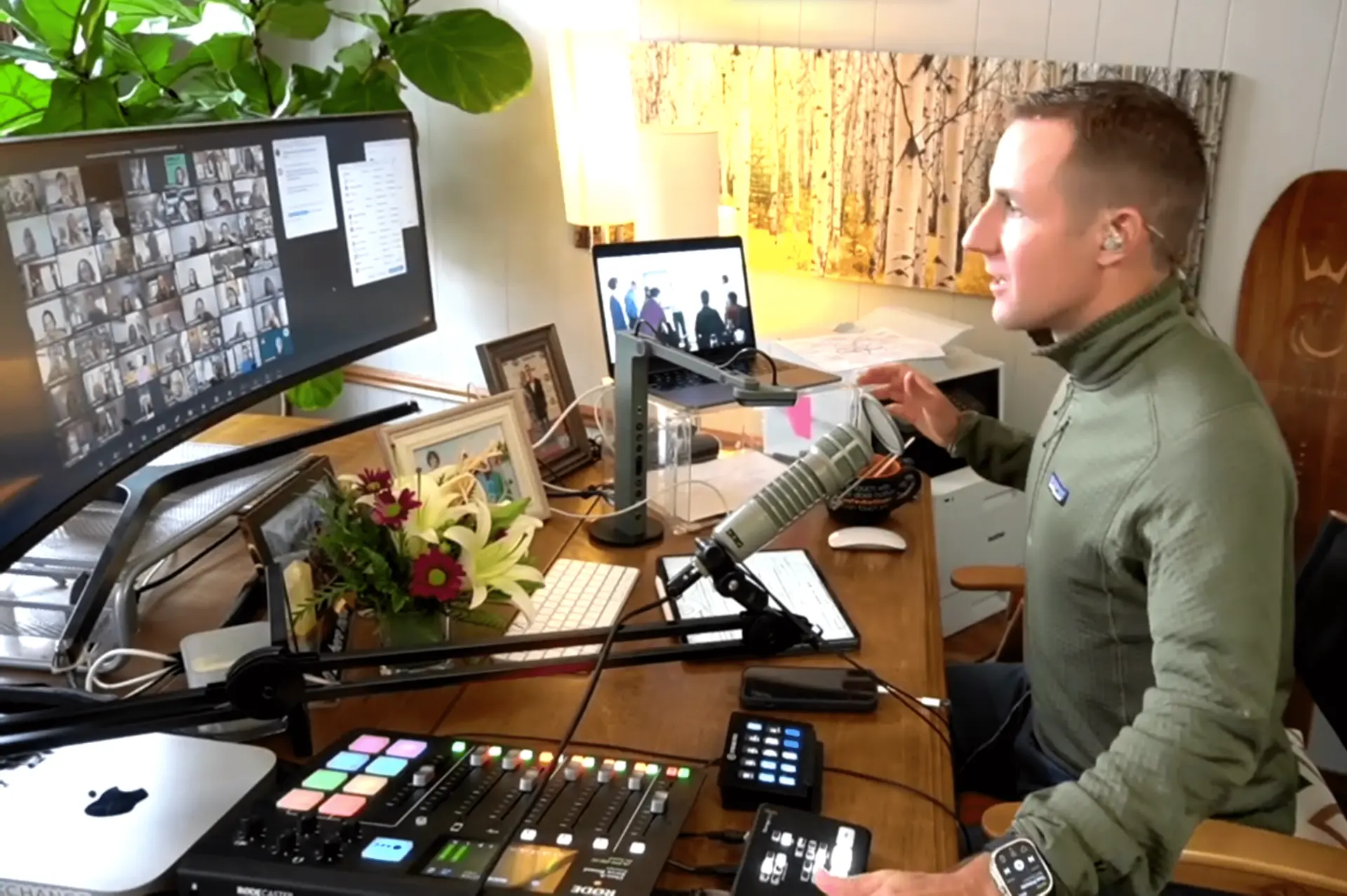 A man wearing an earpiece sits at a desk with audio equipment, a microphone, and a computer. He is on a virtual meeting shown on two screens. The desk has a bouquet of flowers and framed photos. Jon facilitating workshop