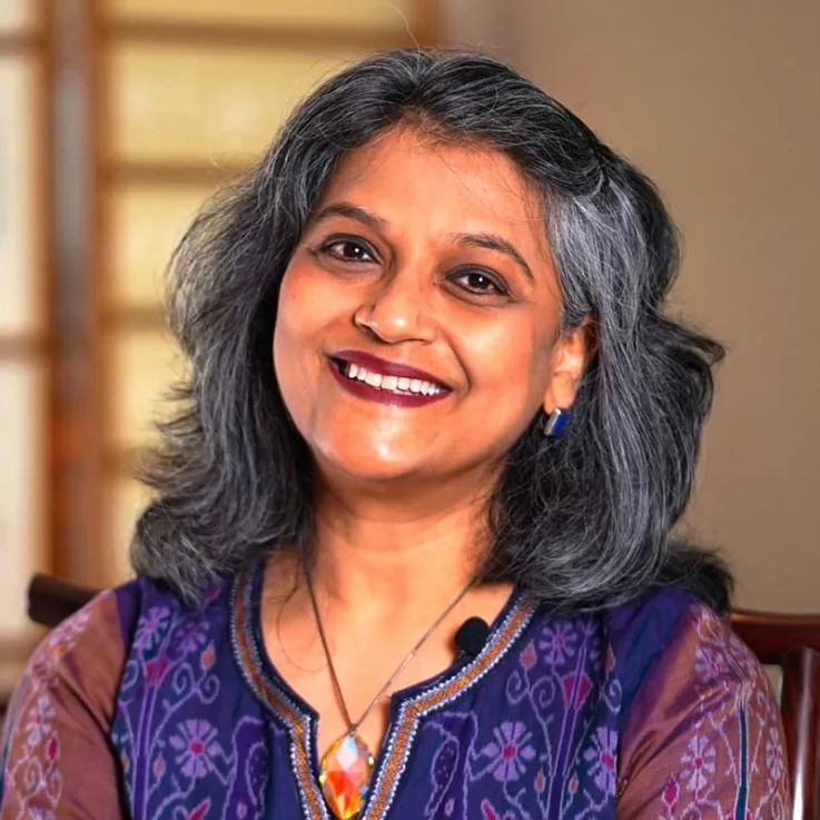 A woman with gray and black hair is smiling at the camera. She is wearing a patterned purple top, a necklace with a large pendant, small earrings, and a microphone clipped to her outfit. The background is out of focus.