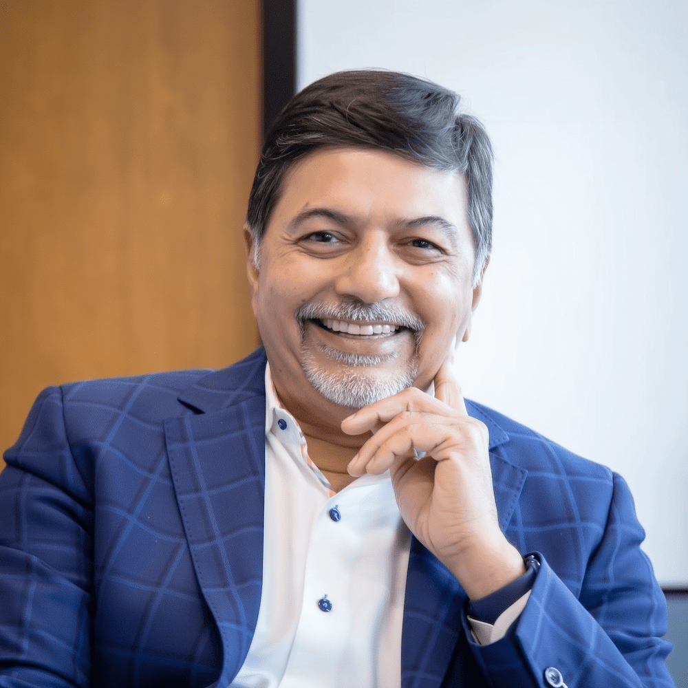A middle-aged man with gray hair and a goatee, wearing a blue plaid suit jacket and white shirt, sits indoors smiling with his hand resting near his chin. The background is out of focus.