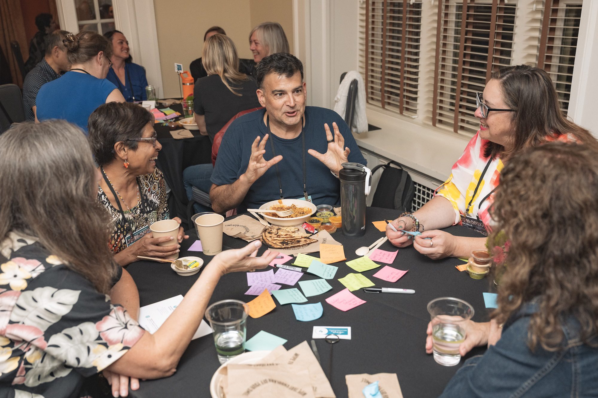 A group of adults sit around a table covered with colorful sticky notes, notebooks, drinks, and snacks, engaged in discussion. One person gestures with hands while others listen and respond.