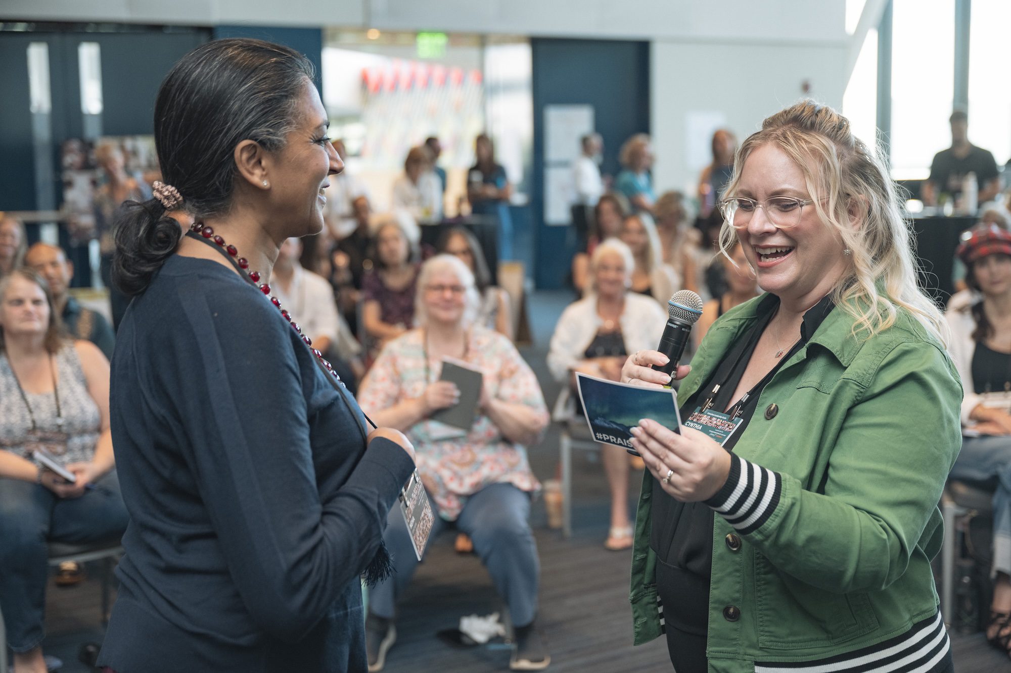 A woman holding a microphone and a card speaks to another woman in front of a seated audience in a bright room. Both women are smiling, and the audience appears engaged and attentive.