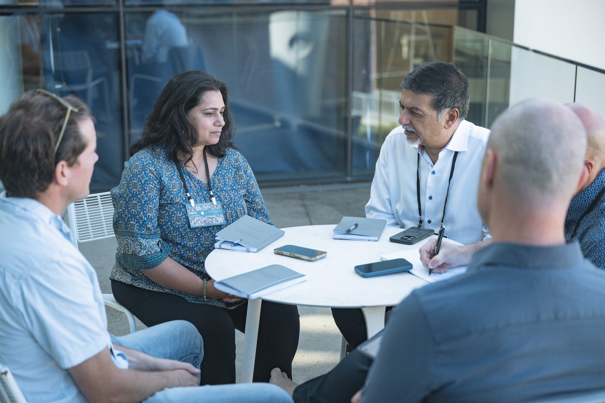 Five people sit around a round table outdoors, engaged in a discussion. Notebooks, a phone, and tablet are on the table. The group includes both men and women, and they appear to be in a professional meeting.