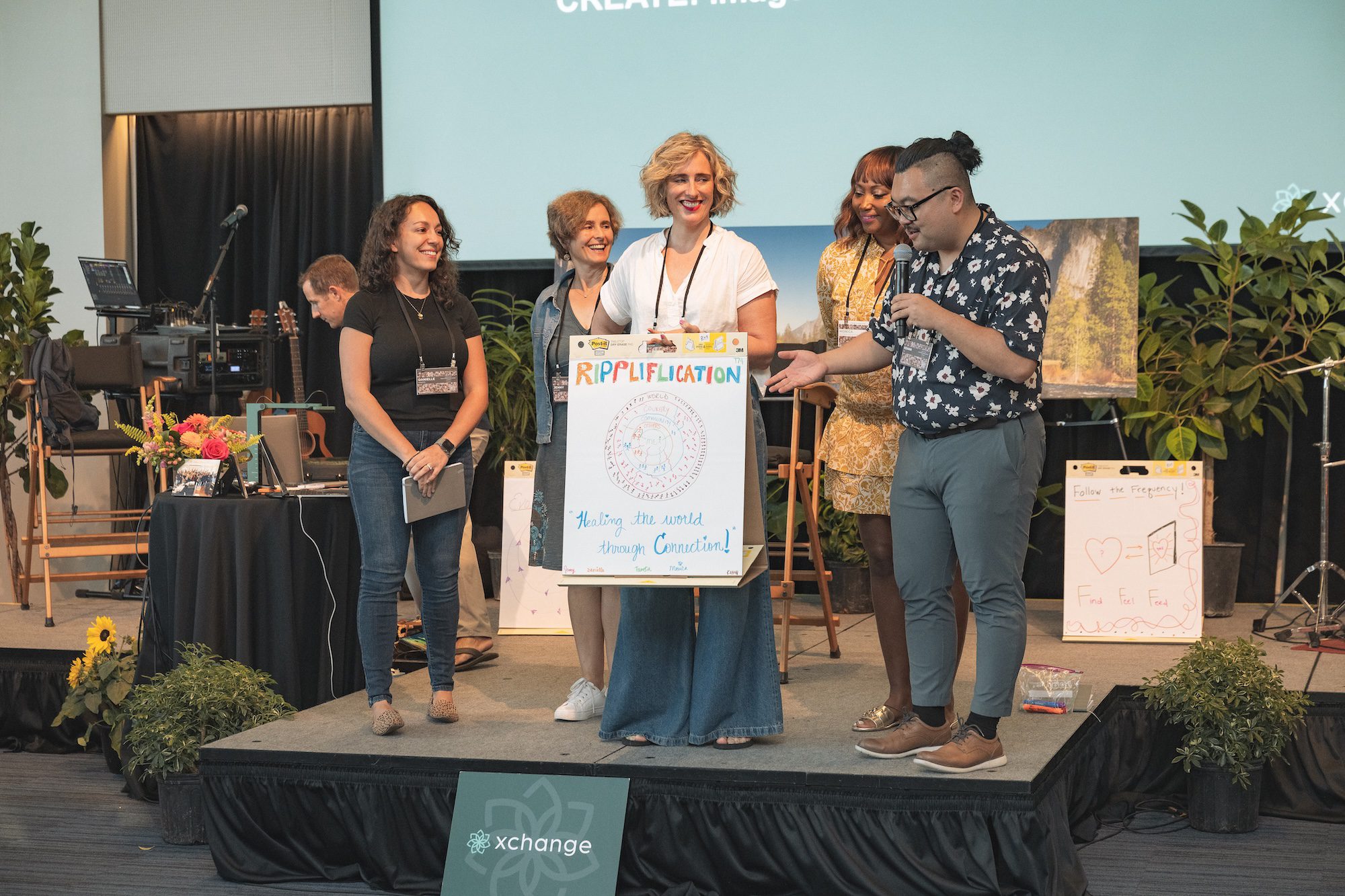 Five people stand on a stage; one person holds a microphone while another holds a large illustrated poster titled "Riplification." Behind them are plants, a black curtain, and presentation materials.