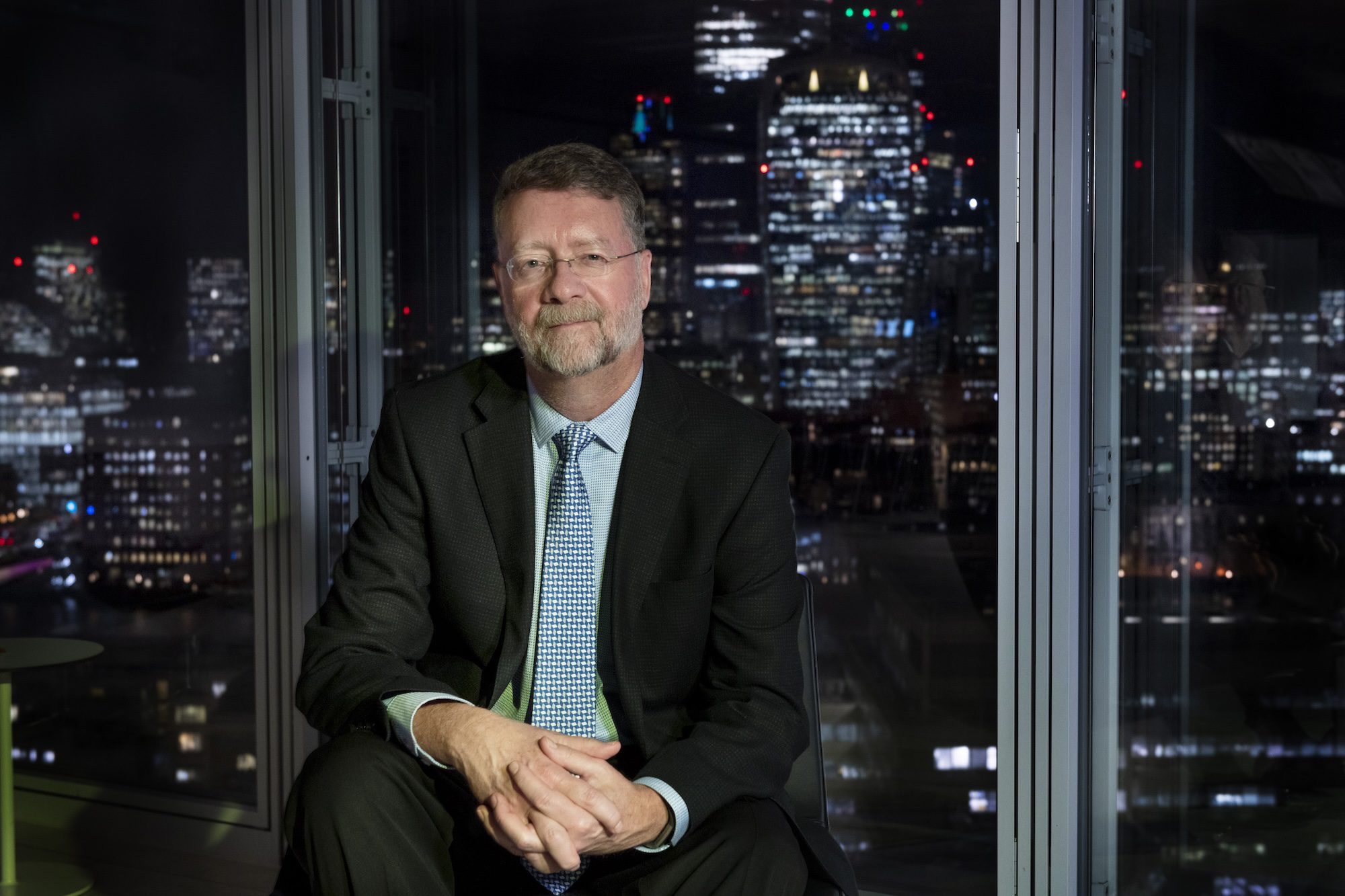 A man with glasses and a beard, dressed in a suit and tie, sits indoors at night with a city skyline of tall, illuminated buildings visible through large windows behind him.