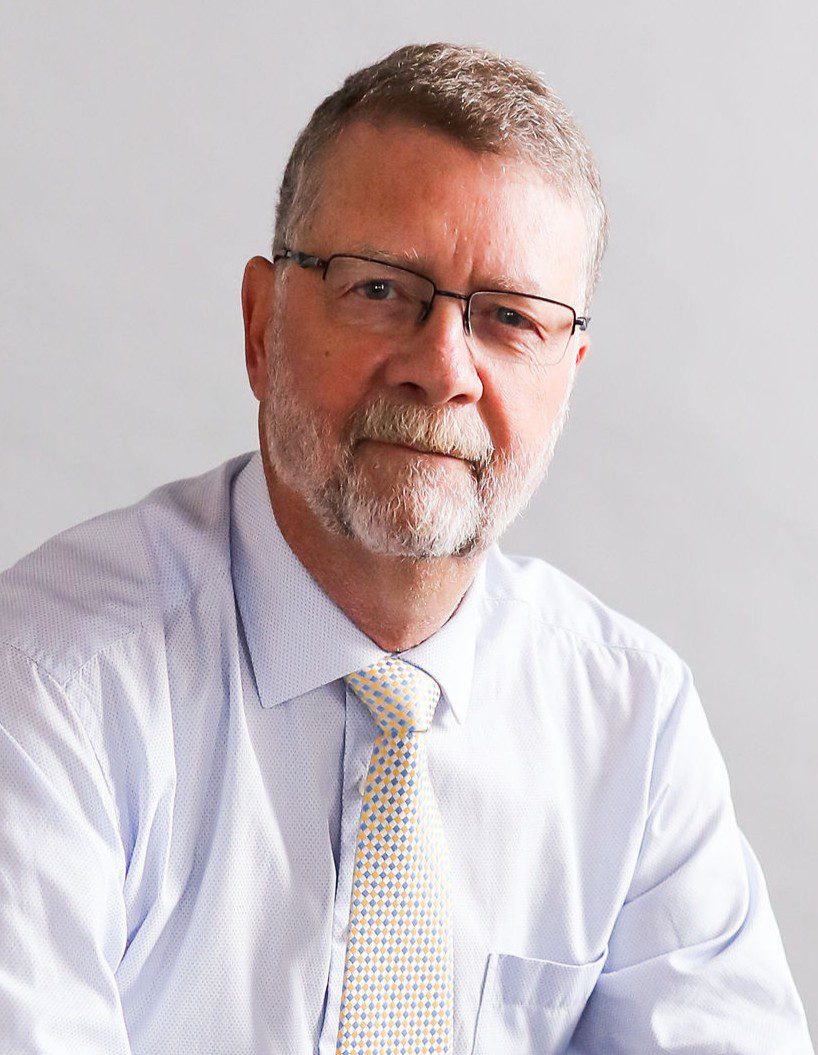 A middle-aged man with short gray hair, a beard, and glasses, wearing a light blue dress shirt and a yellow patterned tie, sits against a plain light background.