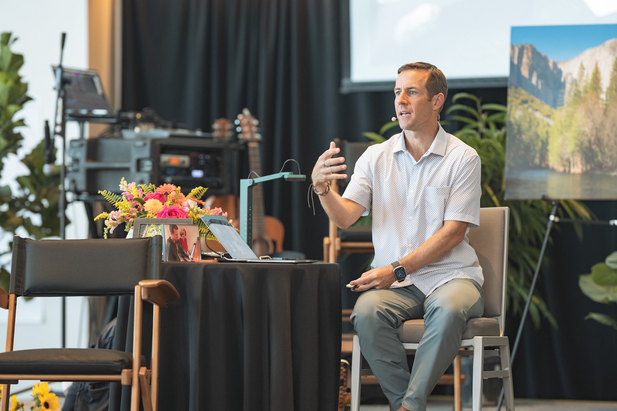 A man in a short-sleeve shirt and slacks sits on a chair, speaking with a gesture. He is next to a table with flowers, a framed photo, and papers. A large nature photo is displayed on an easel in the background.