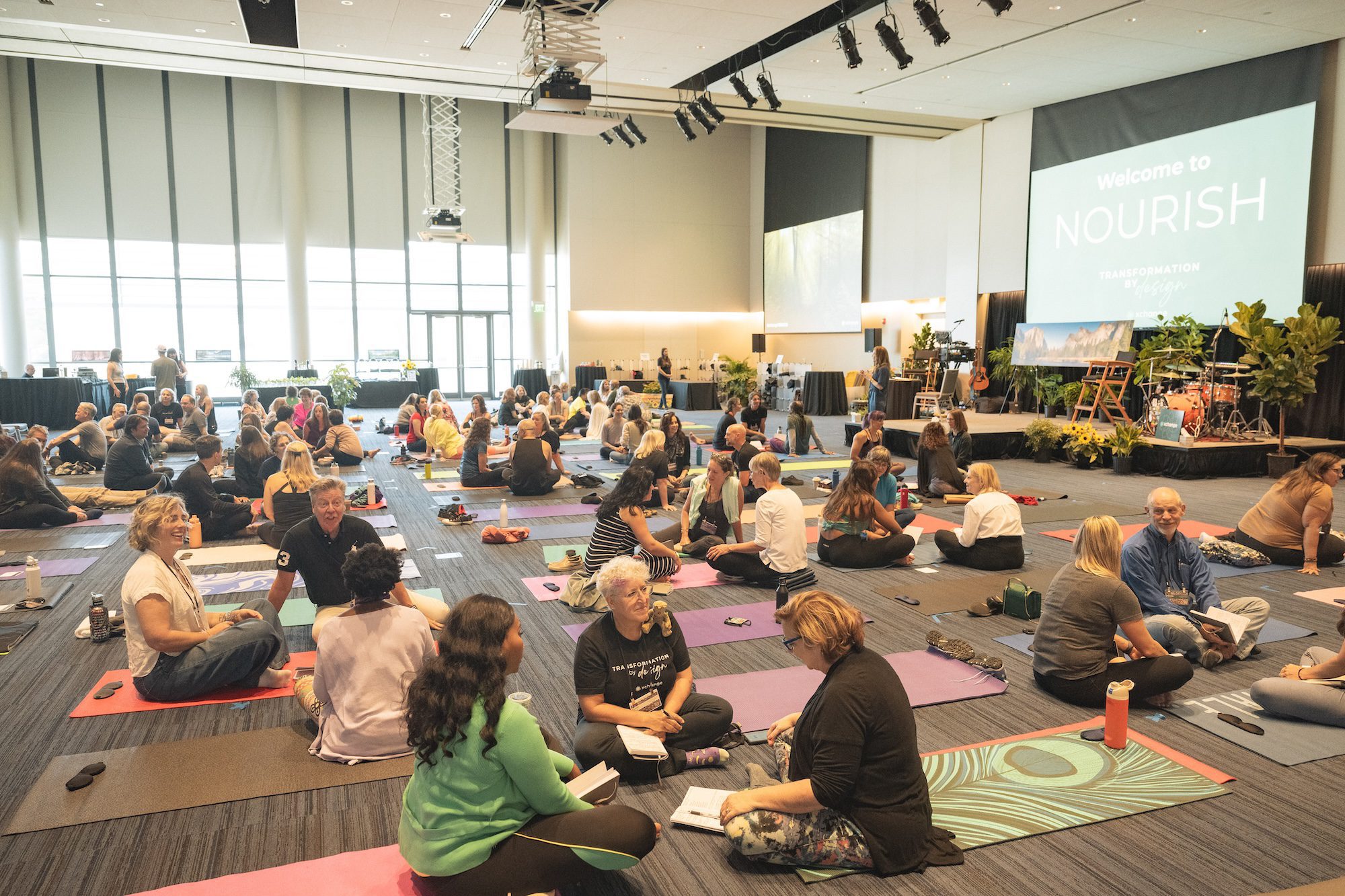 A large group of people sit on yoga mats in pairs or small groups inside a spacious, well-lit conference room. A screen at the front displays "Welcome to NOURISH." Tables and plants decorate the room.