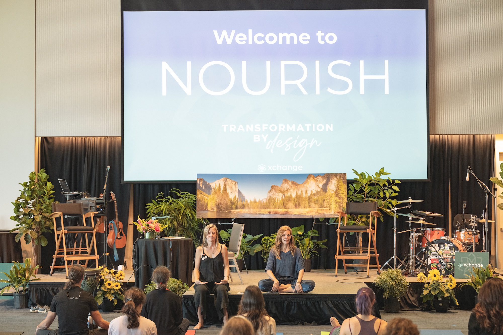 A group of people sit on the floor facing a stage with two women seated cross-legged in front of a large screen that reads "Welcome to NOURISH: TRANSFORMATION BY DESIGN." Musical instruments and plants are on the stage.