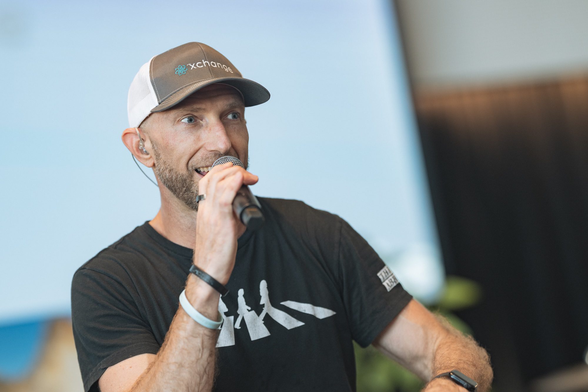 A man wearing a black t-shirt and a gray and white cap speaks into a microphone. He has a beard, is wearing a wristband, and stands indoors with a blurred background.
