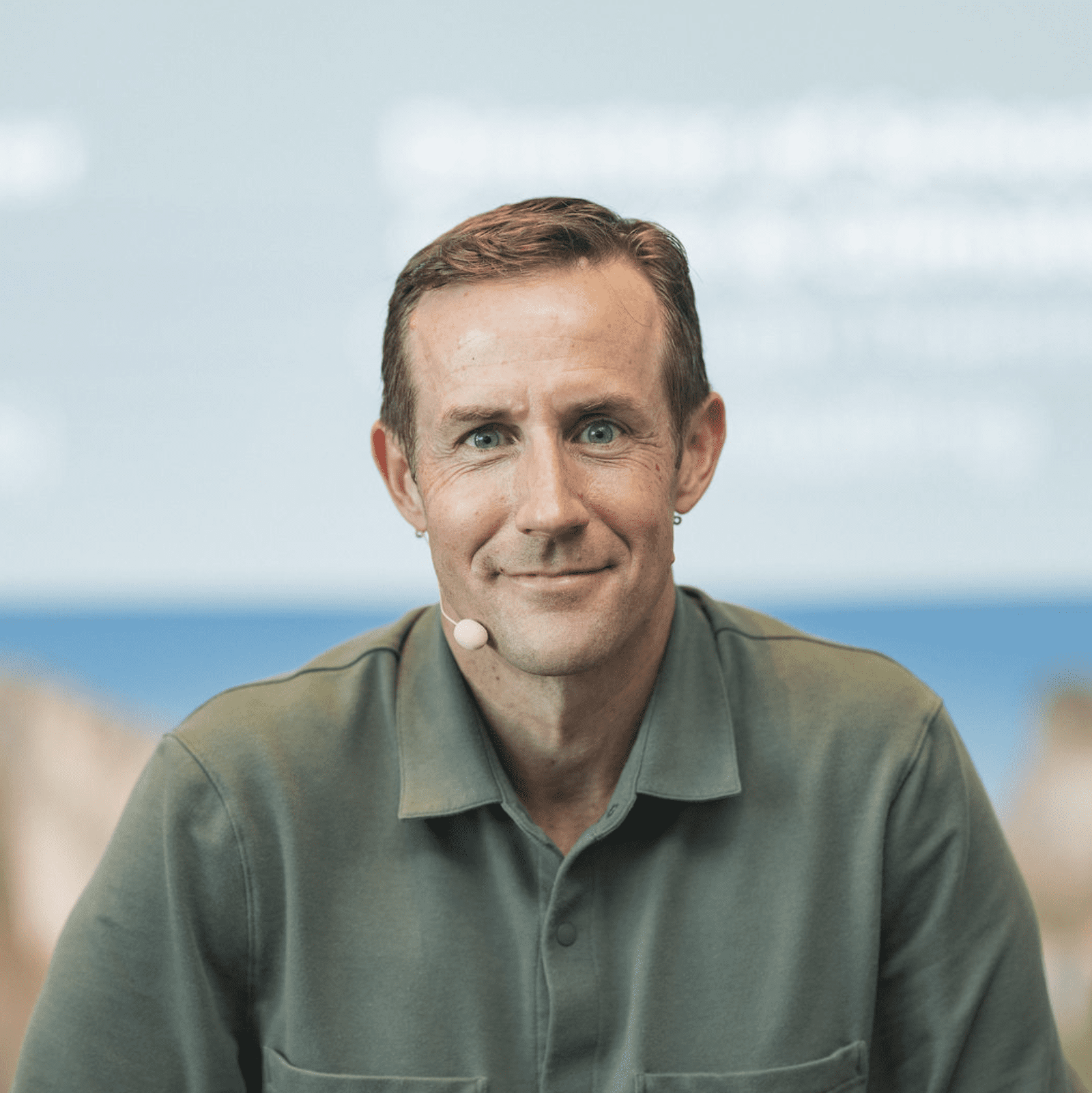 A man with short brown hair, wearing a green collared shirt and a headset microphone, sits in front of a blurred blue and white background. He is facing the camera and smiling slightly.