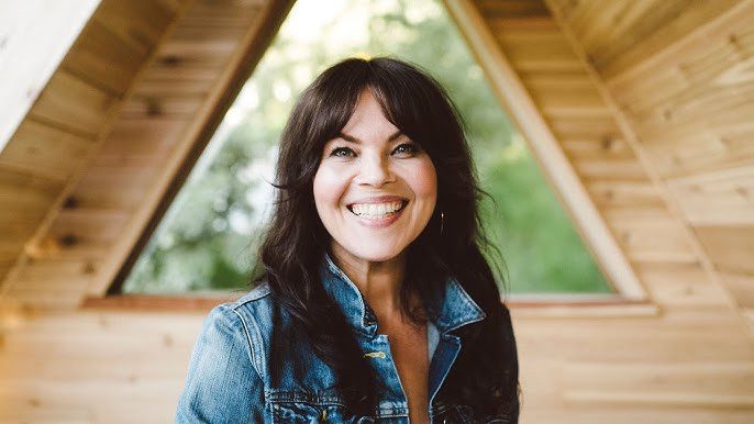 A woman with long dark hair and a denim jacket smiles at the camera while standing inside a wooden A-frame structure with a triangular window in the background.