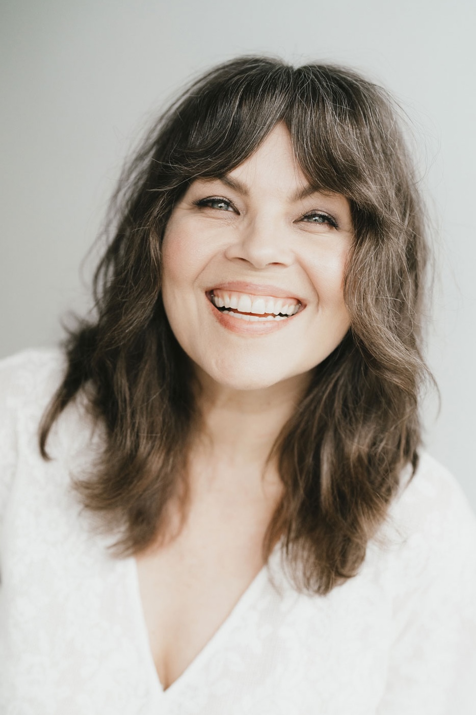 A woman with shoulder-length brown hair and bangs smiles broadly while looking at the camera. She is wearing a white top and is set against a plain, light-colored background.