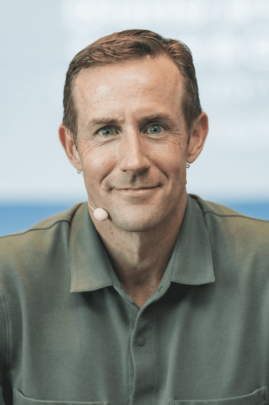A man with short brown hair and a neutral expression wears a green collared shirt and a microphone headset. The background is out of focus and blue-toned.