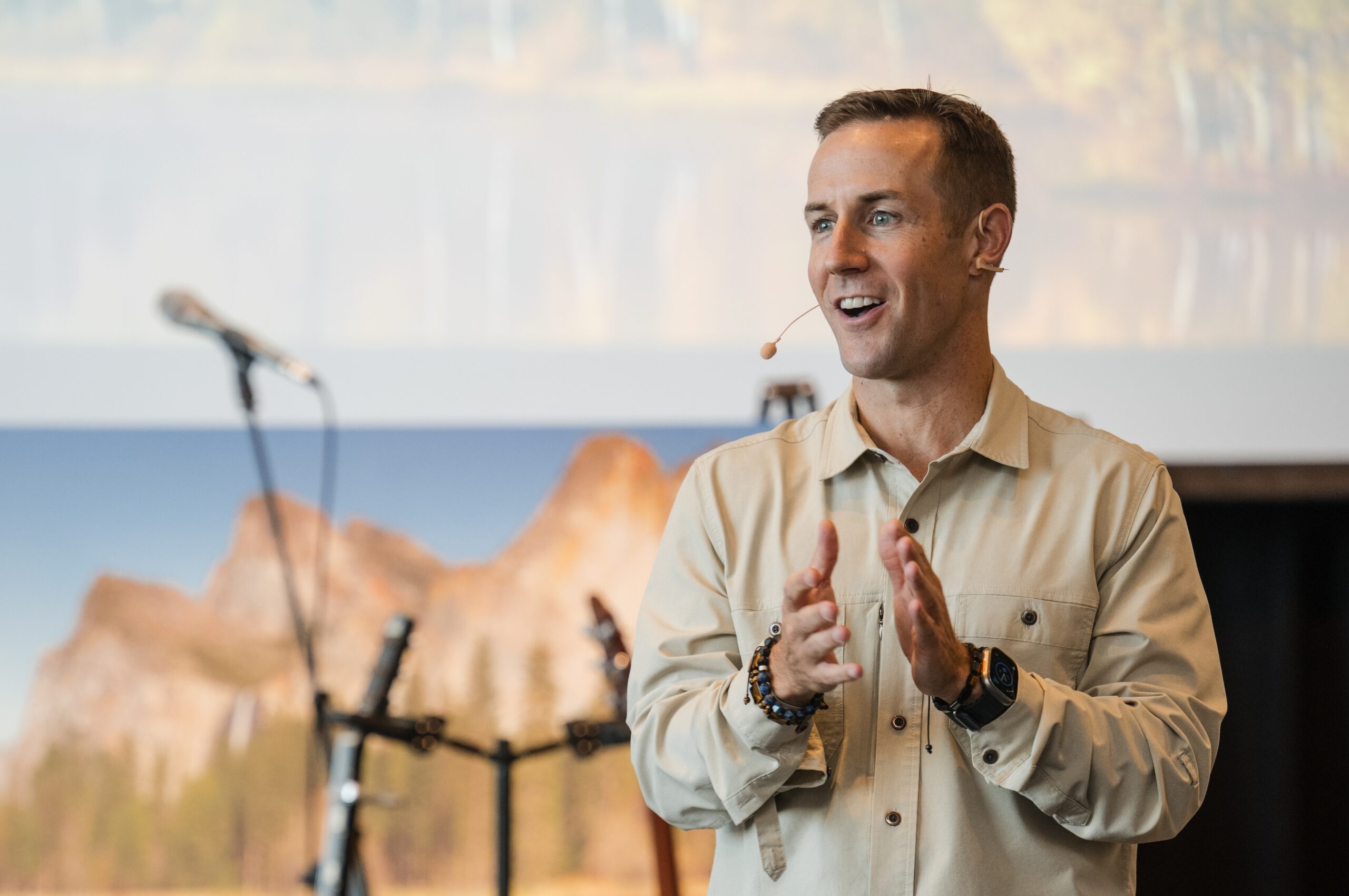 A man wearing a beige shirt and a headset microphone stands and gestures while speaking; microphones and a blurred mountain landscape are visible in the background.