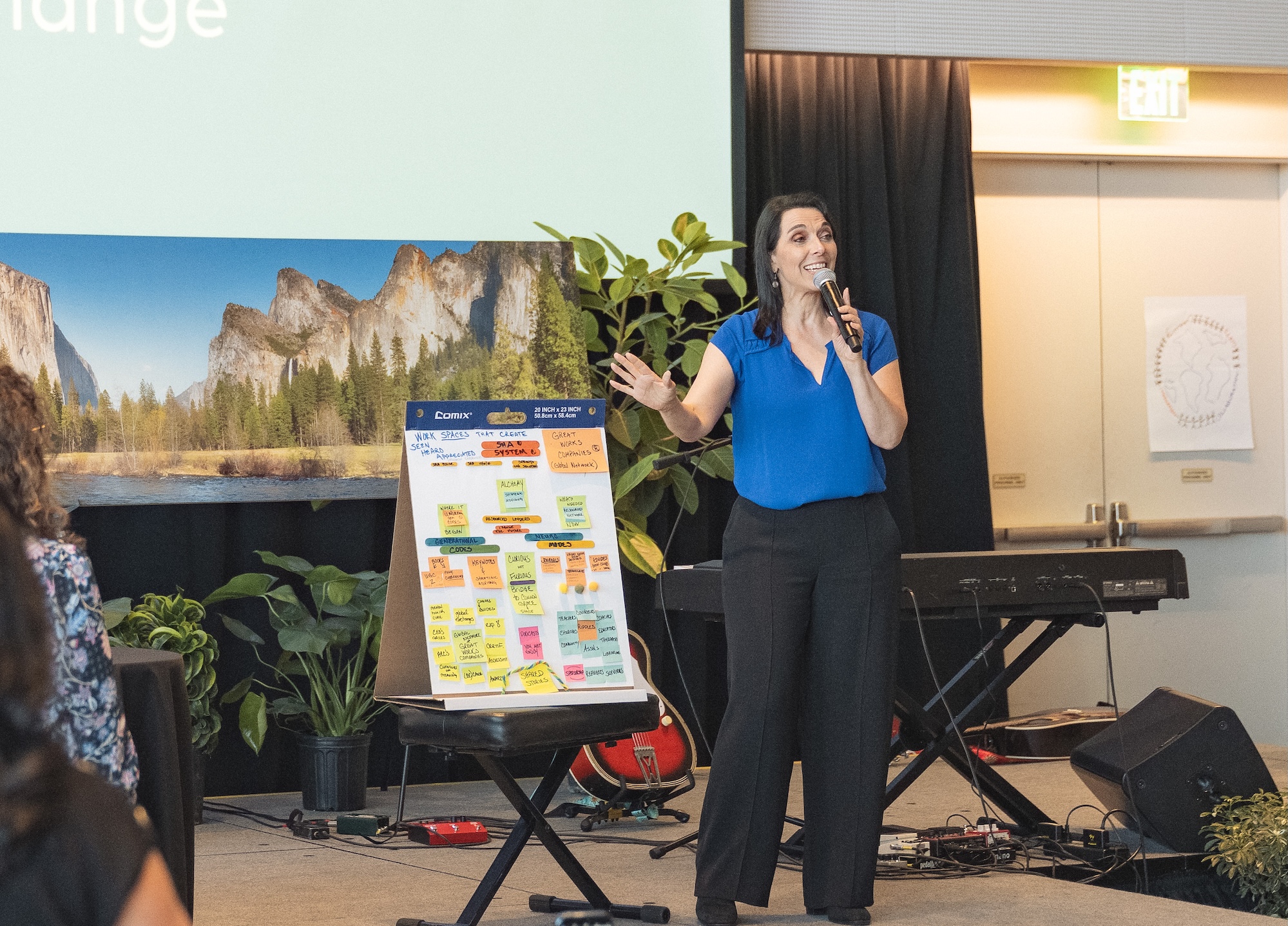 A woman in a blue shirt speaks into a microphone, gesturing toward a flip chart with colorful sticky notes. She stands on a stage with plants, a keyboard, and a Yosemite landscape background. An exit sign is visible.