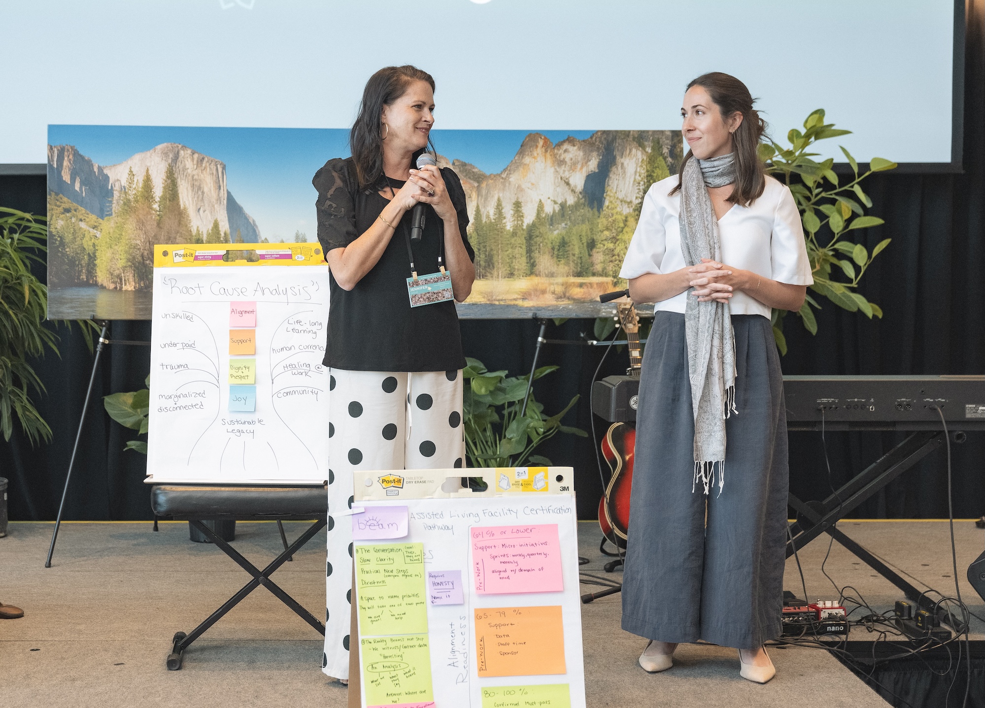 Two women stand on a stage speaking. One holds a microphone. Next to them are easel pads with colorful notes and diagrams. A large scenic photo is displayed behind them, with plants and a keyboard in the background.