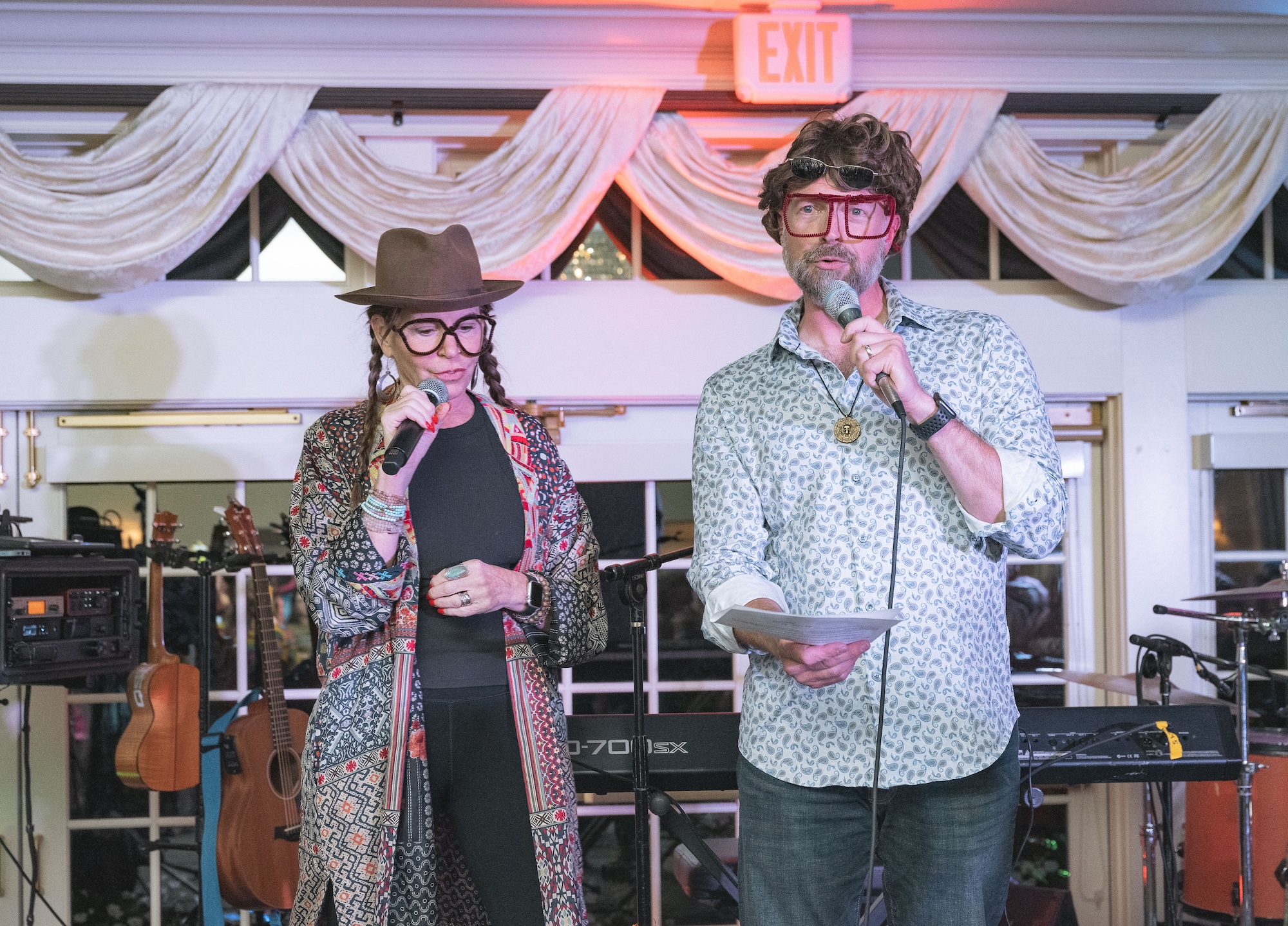 Two people stand indoors holding microphones and papers. Both wear large novelty glasses; one wears a hat and patterned clothing, the other has a button-up shirt and curly hair. Musical instruments are visible in the background.