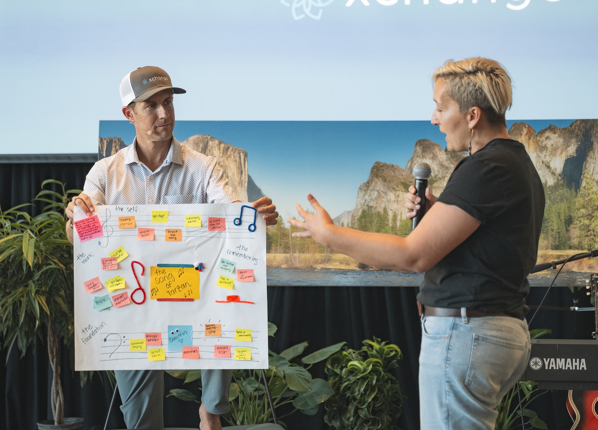 Two people stand indoors; one holds a microphone while the other displays a poster covered with colorful sticky notes and sketches. A nature landscape photo is in the background.