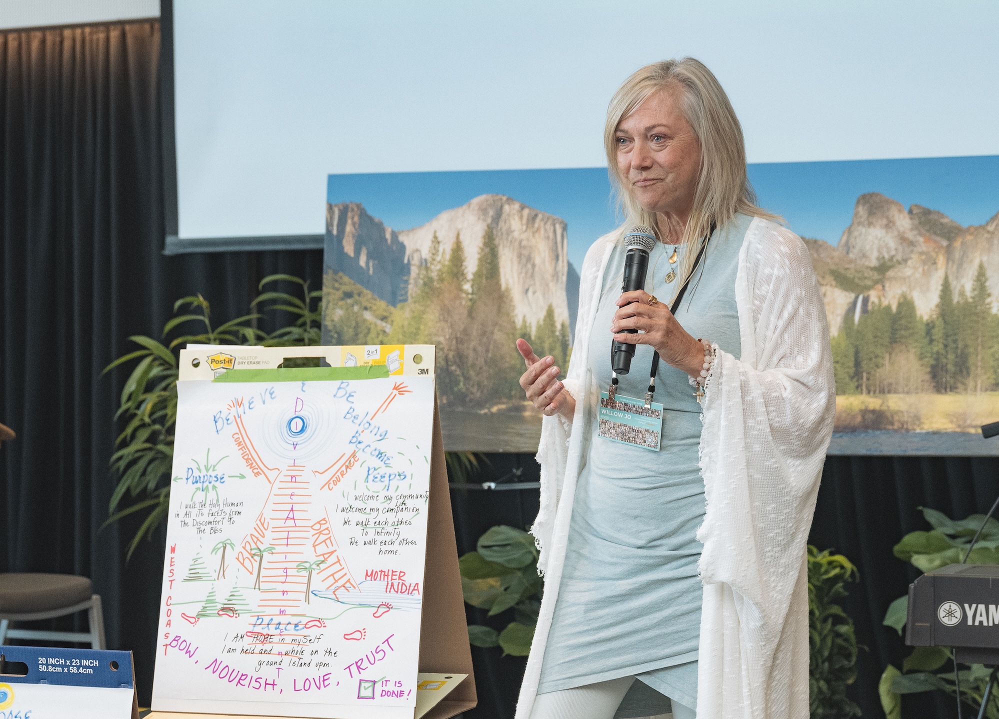 A woman stands holding a microphone next to a flip chart with colorful handwritten notes and drawings. She wears a light blue outfit and white shawl. A nature photo is displayed on a screen behind her.