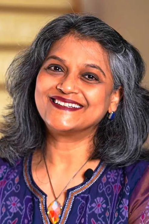 A woman with gray hair and a purple top smiles at the camera. She wears a necklace, earrings, and a microphone on her collar. The background is softly blurred. A woman with gray hair and a purple top smiles at the camera. She wears a necklace, earrings, and a microphone on her collar. The background is softly blurred.
